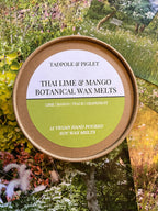 A round Tadpole & Piglet container labeled "Thai Lime & Mango Botanical Wax Melts" rests on colorful garden cards; the label features hand-poured wax melts with lime, mango, peach, and grapefruit scents.