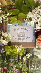 A floral display frames a sign reading "The Biennial Cottage Garden Flower Box" from Bishy Barnabees Cottage Garden, showcasing heritage biennials and an illustration of a house among vibrant flowers and greenery.