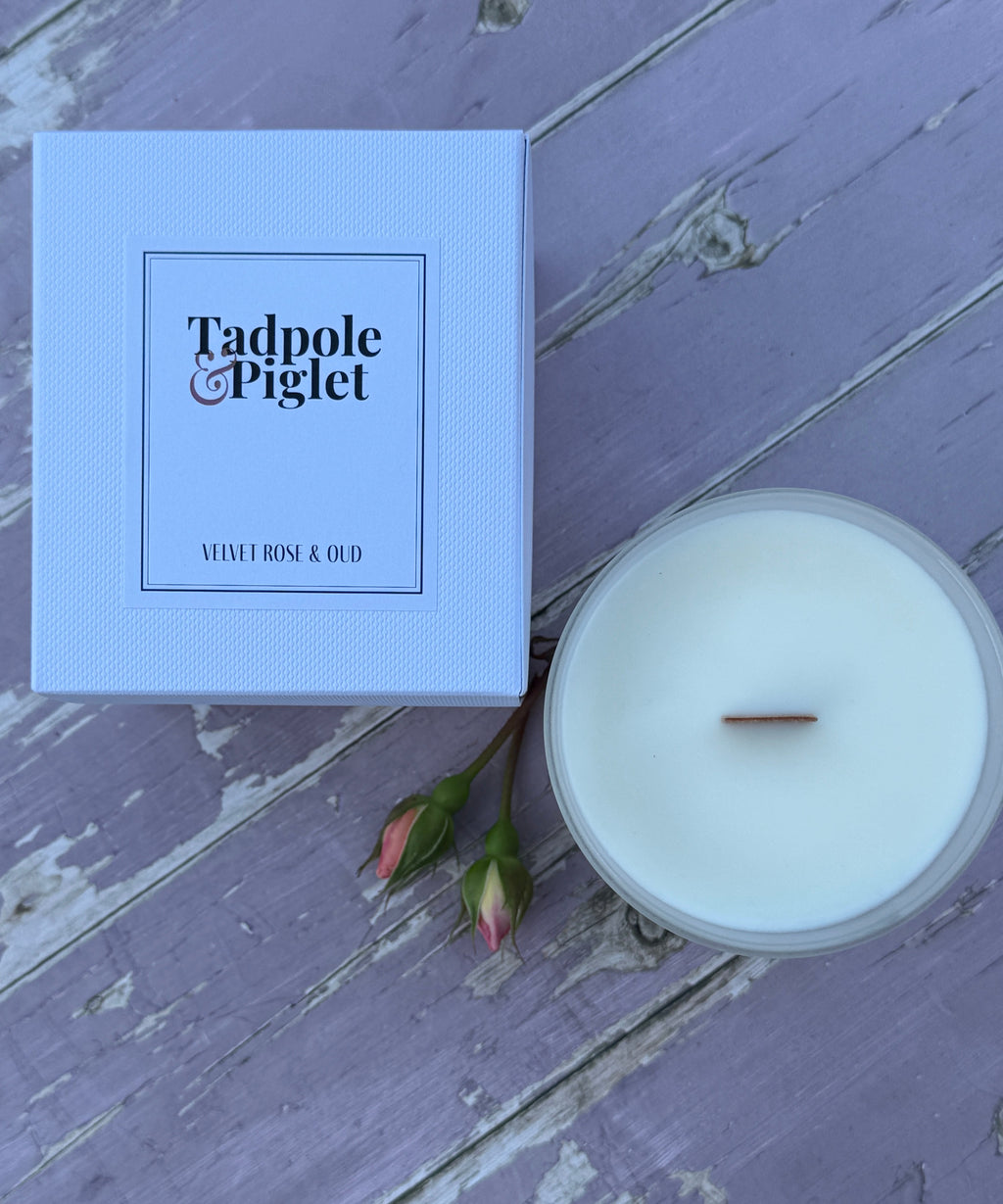 A Tadpole & Piglet Velvet Rose & Oud box sits beside a Velvet Rose & Oud scented candle with a wooden wick and two rosebuds on a rustic purple surface.