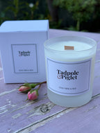 The Tadpole & Piglet Velvet Rose & Oud Scented Candle, made from natural soy wax in a frosted glass jar, sits on a pink wooden surface beside its box and two pink rosebuds, with blurred green foliage in the background.