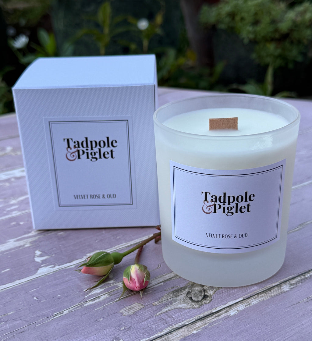 A Tadpole & Piglet Velvet Rose & Oud Scented Candle, made with natural soy wax in a frosted glass jar, rests beside its box on a lavender table with two pink rosebuds and blurred greenery in the background.