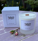 A Tadpole & Piglet Velvet Rose & Oud Scented Candle, made with natural soy wax in a frosted glass jar, rests beside its box on a lavender table with two pink rosebuds and blurred greenery in the background.