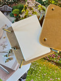 The Bishy Barnabees Cottage Garden Wooden Flower Press - Mini, with bolts, screws, and blank sheets, is displayed atop printed photos of flowers and gardens.