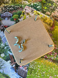 A Bishy Barnabees Cottage Garden Wooden Flower Press - Mini, a square wood piece with four corner screws and metal brackets topped with a nut, is pictured against colorful garden and flower photos.